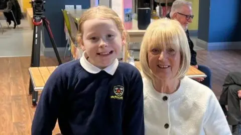 Lynne Aldersley, who is wearing a white cardigan, is sitting on a chair next to Skyla, who is standing. Skyla is wearing her school uniform which includes a dark blue jumper and white shirt. They are both smiling at the camera. 