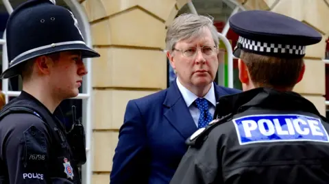 Richard Smith/BBC Roger Hirst is wearing a navy suit and blue tie while talking with two male uniformed police officers. He has grey hair and is wearing glasses.