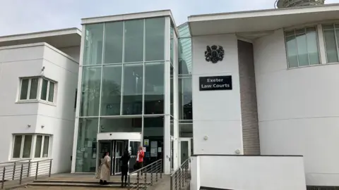 A white crown court building with a large glass entrance. A sign reads Exeter Law Courts. People stand on the steps.