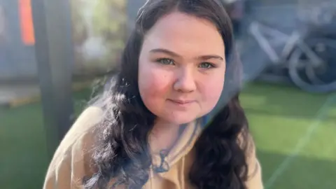 Charly Bisset - A young girl with long wavy dark hair sits outdoors in bright sunlight, wearing a light-coloured hoodie. A bicycle and greenery are visible in the background.
