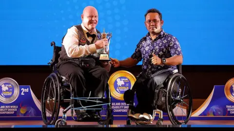 Gary Swift, who uses a wheelchair, dressed in a waistcoat and bow-tie at the World Disability Snooker Championship. He's holding his trophy and is joined by Maitree Kongruang - President of the Sports Association for the Disabled of Thailand.
