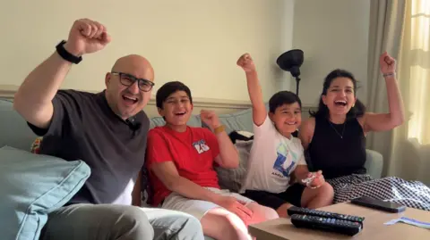 Akmal is wearing a dark grey top and wearing black square glasses, Gafur is wearing a white t-shirt, Bobur is wearing a red t-shirt and Gavhar is wearing a black vested top and a black and white checkered skirt and sitting on a blue sofa. They are all raising their hands saying "Go Ubekistan!". 