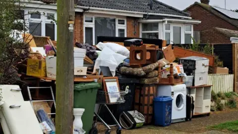 Piles of items including washing machines and furniture are piled up outside the property, which appears to be a bungalow.