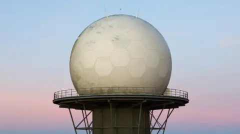 Julian Cartwright A close up of a huge golf-ball like radar on top of a large platform with a fence around its perimeter. The dome is white and has hexagonal shapes on its outside, with small antenna on its top. Behind it is a pink and blue sky