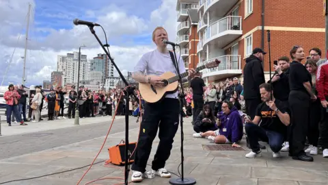 Shaun Whitmore/BBC A man with shaved red hair wearing a white T-shirt plays an acoustic guitar and sings into a microphone. There is a crowd of people around him. Some buildings are in the distance as well as boats on a marina.