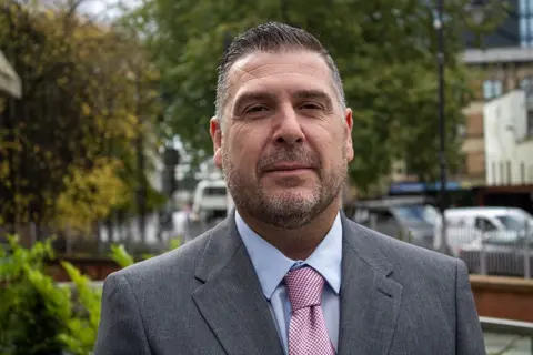 A man in a grey suit, blue shirt and light pink tie, is standing in front of some bushes, with a road with parked cars behind him