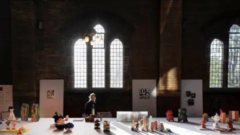 British Ceramics Biennial A large hall inside a church building with various ceramic models displayed on a big white table. A woman can be seen looking at the display.