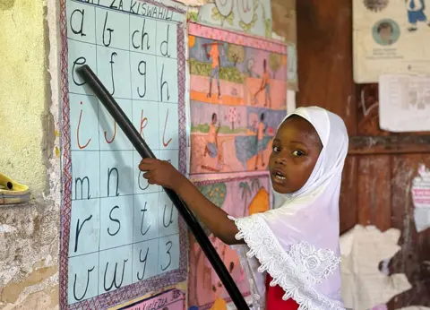 NESE ARI / ANADOLU / GETTY IMAGES A girl reads aloud pointing at the letter on the the wall using a stick.