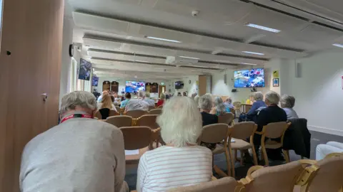 A room with beige-coloured chairs. There are people sitting on the chairs, facing away from the camera. There are TV screens on the walls that show the room. The walls are white and the carpet is grey.