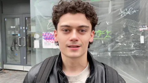 Robby West/BBC Joe Savory. A man with brown curly hair who is looking at the camera and smiling. He is wearing a black leather jacket and is wearing a backpack. He is stood outside. 