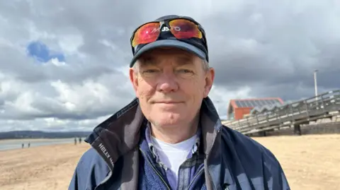 Richard Stokes on the beach wearing a blue jumper and coat. He is wearing a cap with sunglasses on top of it.