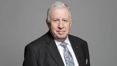 House of Lords An official portrait of Lord Paul Murphy - a man with grey hair wearing a black suit jacket, a white collared shirt and a blue patterned tie smiles at the camera while standing against a grey background.