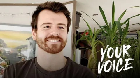 Andrew Hall, a young man with light brown facial hair, standing in his living room smiling, with a painting on the wall and plants seen behind him