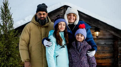 Guz Khan, Morgana Robinson and two girls playing their children, in winter coats and hats in front of a snow-covered log cabin and Christmas tree