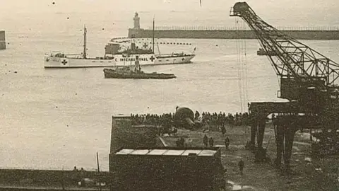 A black and white image of The SS Vega ship in the water in Guernsey's Saint Peter Port harbour in 1944. There is a tug boat nearby and hundreds of people are waiting on the harbour net to a large crane.