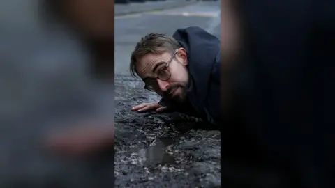 A man in a black coat lies on a road next to a pothole that has water in it.