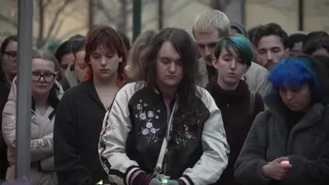 A group of people look down as they stand to observe a moment's silence. Some are holding candles.