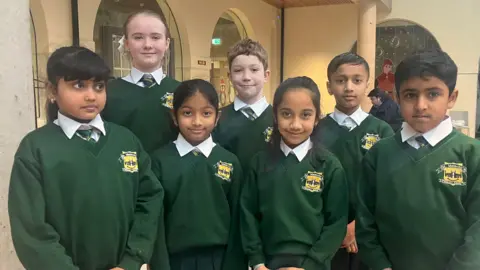 Seven of the Irish language class pupils at St Malachy's Primary School in Belfast standing in two lines.  They are all wearing their school uniform -  dark green school jumpers, white shirts and green and yellow striped ties.