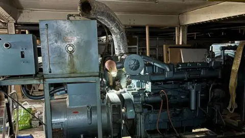 Ukrainian Action The generator is blue and old-looking. There are a number of mechanical parts and one person is working on it with a torchlight. 