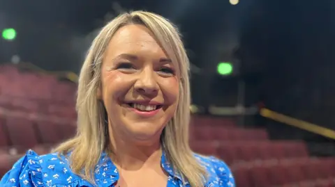 Emma Baugh/BBC A head and shoulders image of Natalie Phillips. She is smiling and looking into the camera. She is standing in what appears to be an auditorium. She is wearing a blue top and there are seats behind her.