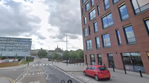 Rotherham Council's Riverside House in the town, a red stone building on the right. A red car is parked outside in a disabled space. A Zebra crossing is ahead, before a bridge over the River Don, with two church spires and trees in the distance. A modern office block is on the left of the road.