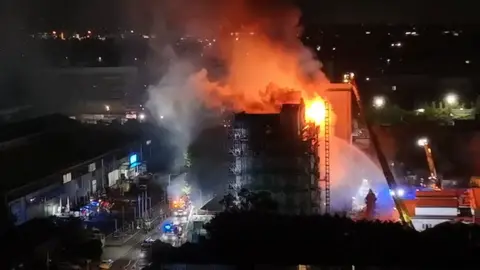A large fire engulfs the upper floors of the Spectrum Building at night, with flames and thick smoke rising into the sky. Fire crews and emergency vehicles are visible below, with water jets being directed at the blaze.