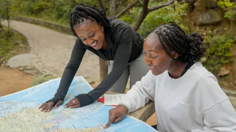 Two young women both looking at a map on a table. One is sitting and pointing at the map while the other looks over her shoulder.