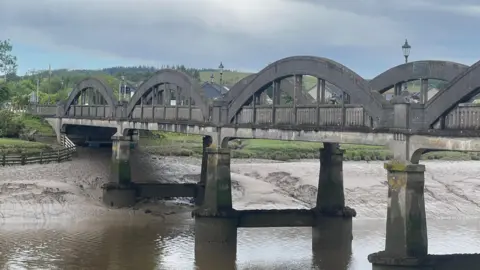 A greying stone bridge crossing the river with a reflection