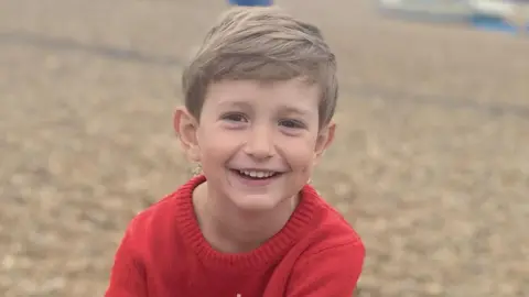 Benedict, a young boy with blond hair, is on a pebbly beach. He is wearing a red knitted top and is smiling to the camera.