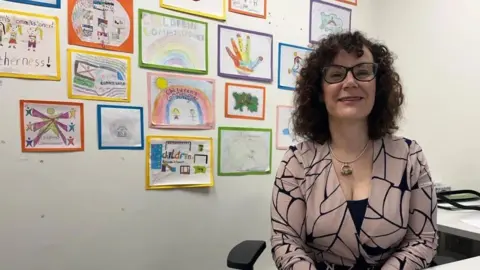 Carmel Corrigan sits at a desk smiling at the camera. On the wall behind her are many colourful pictures of people, rainbows and other scenes that look like they have been drawn by children.