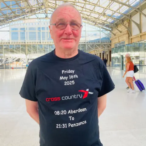 Man - John Labrow - smiling at camera in station, wearing a t-shirt which says 'Friday May 16th 2025 / CrossCountry / 08:20 Aberdeen to 21:31 Penzance'.