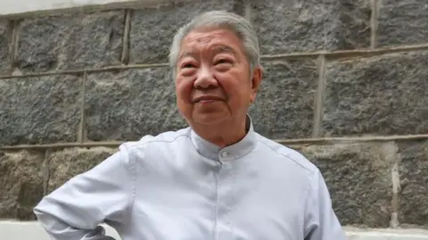 Chua Lam wearing a white button-up shirt, standing in front of a grey wall with his hand on his hip