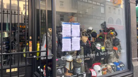 A shop window with four police noticed placed in it.  In the shop are masks and military headwear