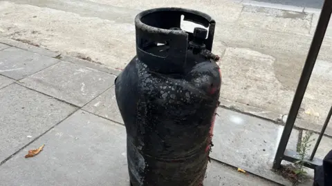 A gas canister, that is mostly black and charred, placed upright on the pavement. There are patches of red showing through the charring. 