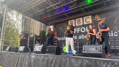 Ollie Ward/BBC A band perform on a festival stage. Several members play instruments including trumpets, saxophones, guitars and drums. There is a man singing into a microphone. 