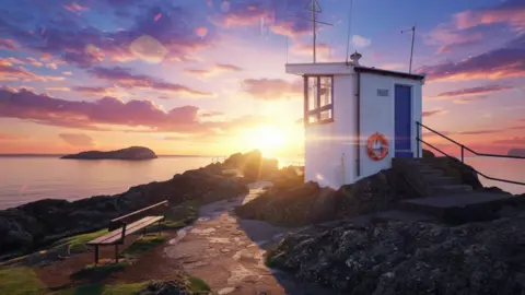 Jacqueline Dawson A small, white harbour lookout building stands on a rocky shoreline at sunset. Bright sunlight and colourful clouds illuminate the scene, with a bench, coastal path, and offshore island visible in the background.
