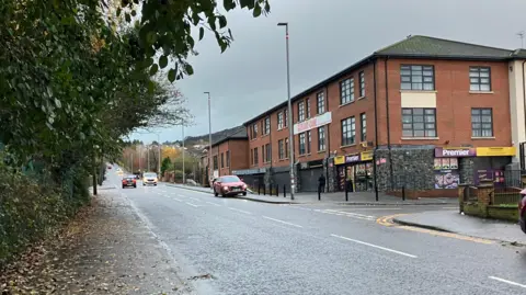 A street with a modern red brick building and shopfronts at the bottom. Ove red car is parked on the pavement and a couple of cars are driving down the street. 