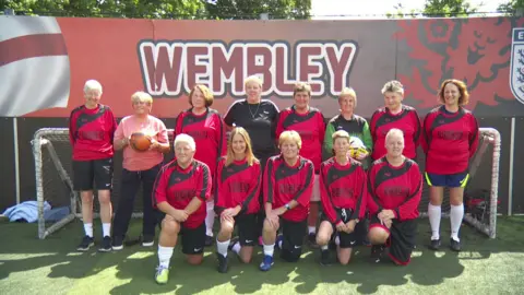 A women's team during a practice. They are smiling for the camera. They are wearing red and black kits. There is a placard reading Wembley behind them.