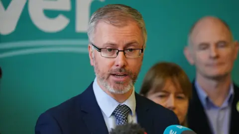 PA Media Roderic O'Gorman in a blue shirt and navy suit jacket against a green wall, speaking into a microphone with team members in the background