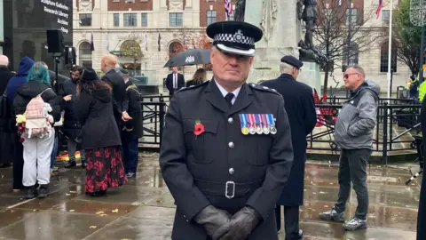 Kaushal Menon Muralidharan/BBC A uniformed police officer stands facing the camera. He is wearing a cap, dark gloves and a dark uniform with several medals on the chest and a red poppy pin. Behind him are a group of people gathered near a tall stone war memorial. The memorial features a bronze statue of a soldier with several Union Jack flags visible.