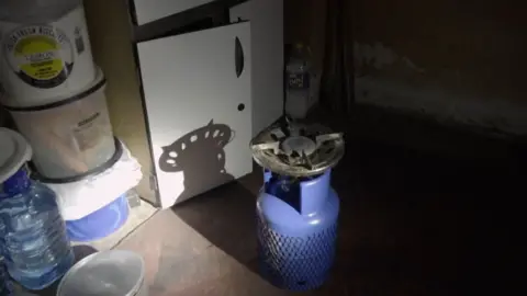 A blue gas canister and stove are illuminated by torchlight. A bottle of water and some other containers can be seen nearby.