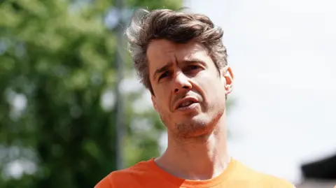 A head and shoulders shot of Dr Patrick Hart wearing an orange t-shirt and looking into the distance