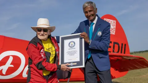 Guinness World Records Harry Heasman, in a red and black jumpsuit and yellow T-shirt, poses with a man in a blue Guinness World Records branded blazer. The pair are holding up a framed certificate in recognition of Heasman's record. Behind them is the tail of a red plane.
