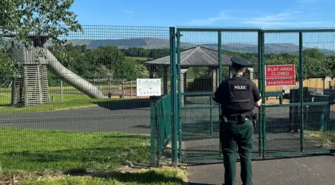PSNI A police officer is seen standing beside a play park in Strathfoyle. The gates are closed.