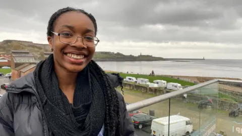 Deborah is standing on a balcony with the River Tyne behind her. She's a teenager with her hair in braids and wearing a black coat and scarf. 