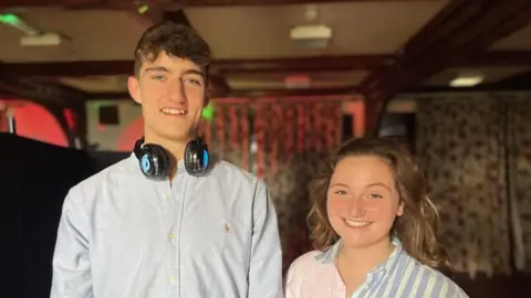 A young boy in his early twenties next to a young women also in her early twenties. The boy is very tall, with brown hair and blue eyes, he is wearing a blue shirt and has big black headphones around his neck. The girl is wearing a stripy blue and pink shirt, she has curly brown hair and is smiling at the camera.