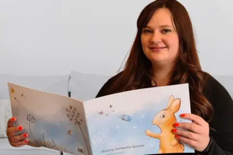 Supplied A woman with brown hair holding up a book called MISSING SOMEONE SPECIAL. It has a light brown rabbit on the front pointing to a butterfly. 