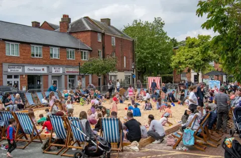 A large number of children and adults sat on a large, rectangular sandy area in a town, with blue striped deck chairs surrounding the sand and red brick buildings in the background