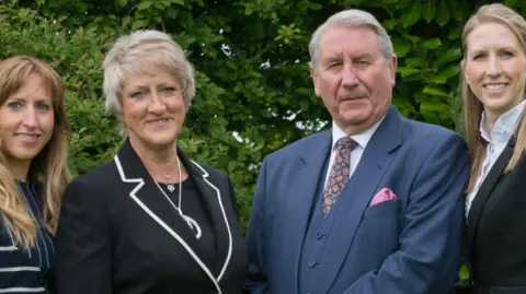 AUBREY KIRKHAM FUNERAL DIRECTORS Aubrey Kirkham pictured in a blue suit with a pink pocket square stood next to wife Ann who wears a black suit and silver jewellery and their eldest daughter Leanne who is also smiling and wearing a blue and white striped shirt.