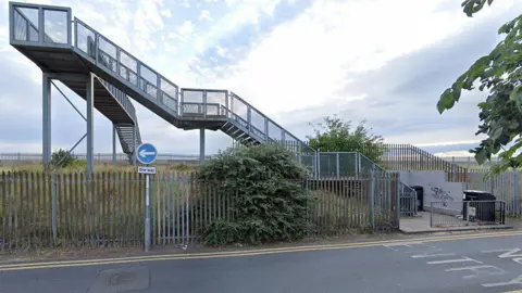 Google street view of the Fuller Street bridge, which is a grey metal structure standing alongside a metal fence with grassland behind it. There is a road in front and a One Way blue sign with a white arrow pointing to the left.